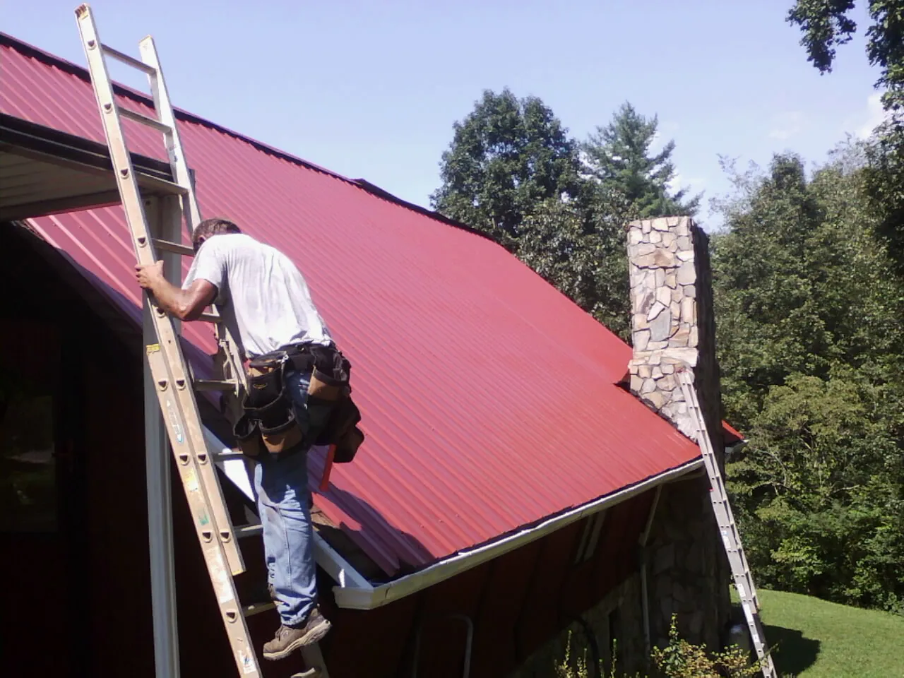 Roofer on a ladder beside a red metal roof and stone chimney — Soffit & Fascia Repair by Roofing Brothers in Orient